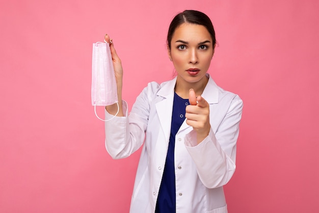 Portrait d'une jeune femme médecin en blouse blanche tenant un masque médical debout isolé sur fond rose et pointant le doigt vers la caméra avec des émotions négatives.