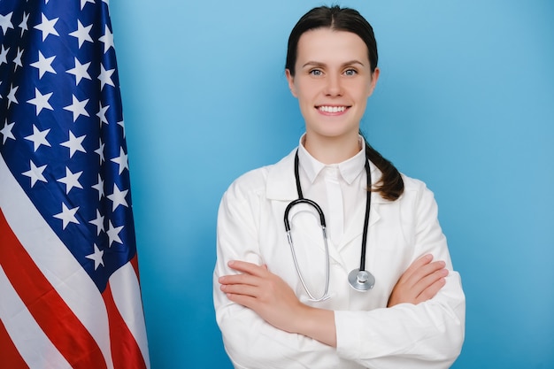 Photo portrait d'une jeune femme médecin heureuse en uniforme blanc et stéthoscope debout près du drapeau national des états-unis, poitrine croisée, posant sur fond bleu. concept de système de santé national aux états-unis