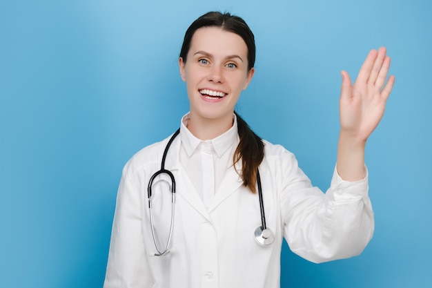 Photo portrait d'une jeune femme médecin joyeuse vêtue d'une blouse blanche médicale et d'un stéthoscope saluant disant bonjour heureux et souriant, geste de bienvenue amical, posant isolé sur un mur de fond bleu studio
