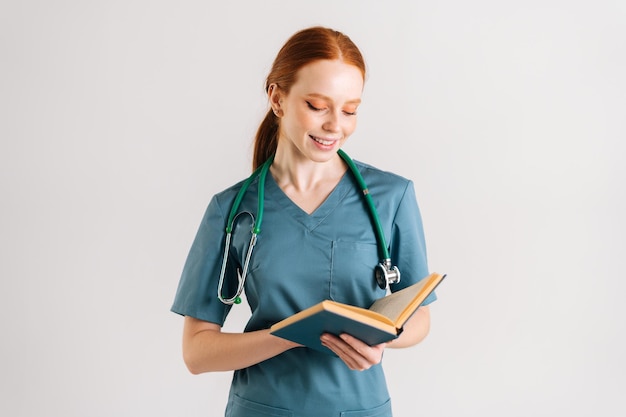 Portrait d'une jeune femme médecin positive en uniforme vert avec stéthoscope lisant un livre médical debout sur fond blanc isolé en studio Femme médecin lisant un traité papier