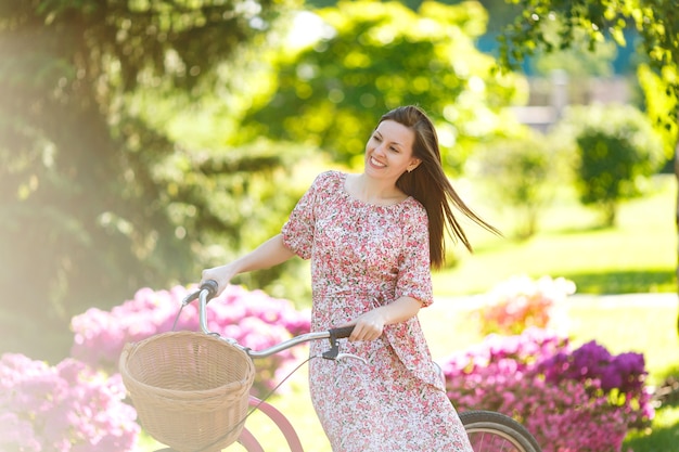 Portrait d'une jeune femme à la mode en longue robe à fleurs rose s'arrêtant pour faire du vélo vintage avec panier pour les achats sur fond de fleurs à l'extérieur. Temps de récréation assez féminin au printemps ou au parc d'été