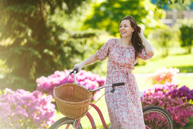 Portrait d'une jeune femme à la mode en longue robe à fleurs rose s'arrêtant pour faire du vélo vintage avec panier pour les achats sur fond de fleurs à l'extérieur. Temps de récréation assez féminin au printemps ou au parc d'été