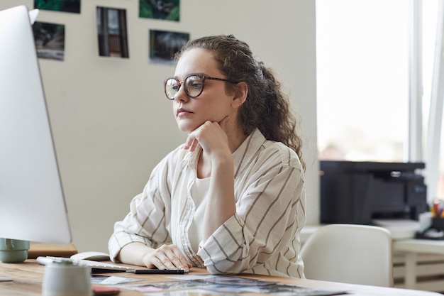 Portrait de jeune femme moderne portant des lunettes regardant l'écran de l'ordinateur pensivement tout en travaillant au bureau dans le bureau blanc, copiez l'espace