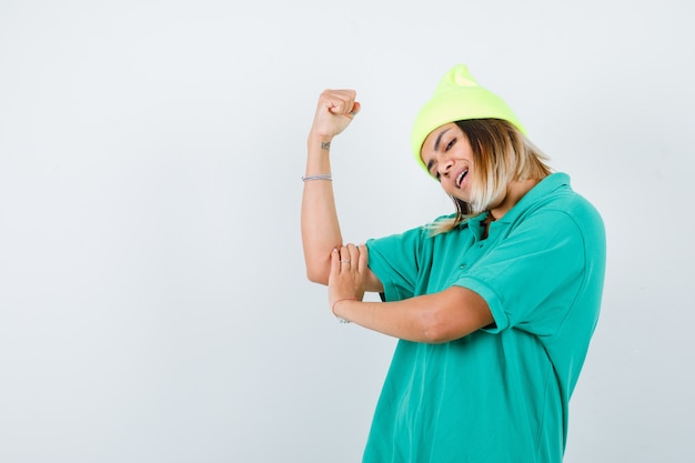 Portrait de jeune femme montrant les muscles des bras en t-shirt polo, bonnet et à la vue de face joyeuse