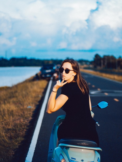 Photo portrait d'une jeune femme montrant un signe de paix assise sur un scooter à moteur sur la route