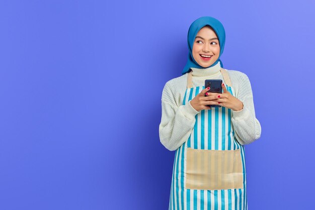 Portrait d'une jeune femme musulmane asiatique souriante de 20 ans portant le hijab et un tablier tapant des messages sms sur un téléphone portable tout en regardant de côté isolé sur fond violet. Concept de mode de vie musulman femme au foyer personnes