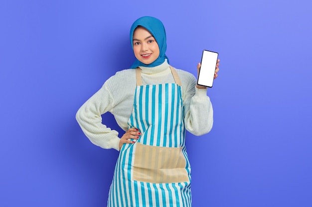 Portrait d'une jeune femme musulmane asiatique souriante portant le hijab et un tablier tenant un téléphone portable à écran blanc isolé sur fond violet. Concept de mode de vie musulman femme au foyer personnes