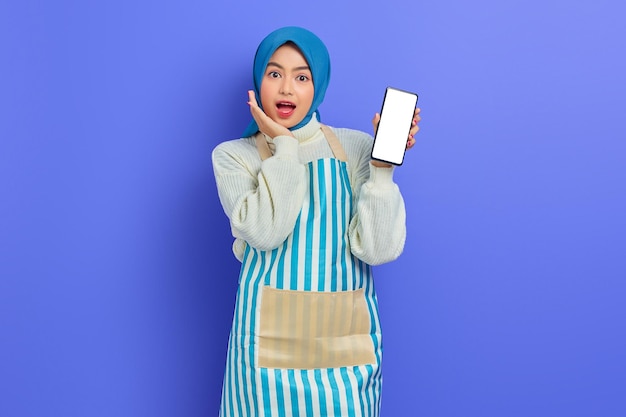 Portrait d'une jeune femme musulmane asiatique surprise portant le hijab et un tablier montrant un téléphone portable à écran blanc isolé sur fond violet. Concept de mode de vie musulman femme au foyer personnes