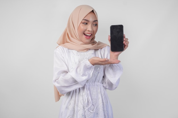 Photo portrait d'une jeune femme musulmane asiatique vêtue d'une robe blanche et d'un hijab souriante heureuse pointant vers l'espace de copie d'écran vide sur son téléphone et montrant l'écran à la caméra
