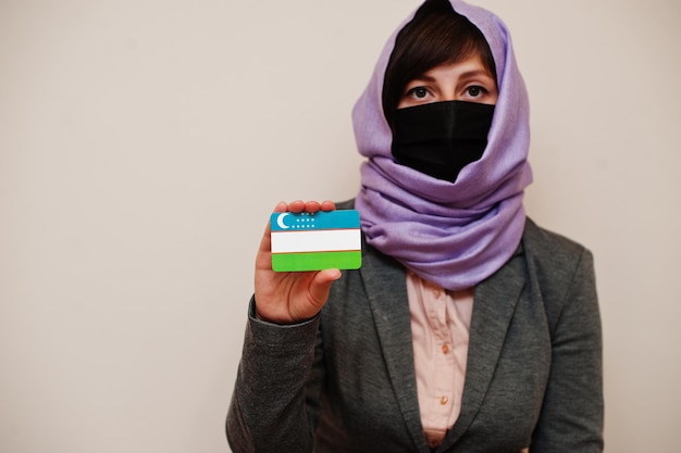 Portrait d'une jeune femme musulmane portant des vêtements de cérémonie protégeant le masque facial et le foulard hijab tenant la carte du drapeau de l'Ouzbékistan sur fond isolé Concept de pays Coronavirus