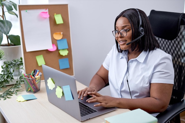 Portrait d'une jeune femme noire agréable représentante du service client avec casque au bureau, belle dame en chemise blanche formelle engagée dans le travail, dans un bureau confortable et lumineux, utiliser un ordinateur portable moderne