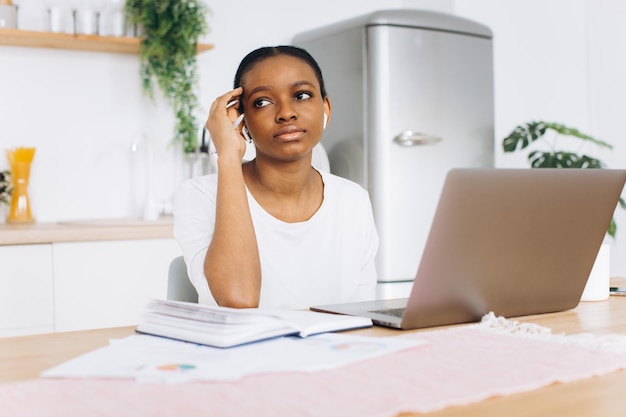 Portrait d'une jeune femme noire assise dans la cuisine et travaillant sur un ordinateur portable à la maison