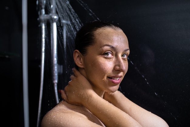 Portrait de jeune femme nue souriante bénéficiant d'eau qui coule, prendre une douche, debout dans la salle de bain tenant les mains sur le cou en prenant soin de sa peau sur le mur noir