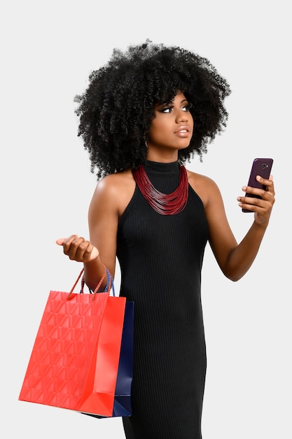 Portrait d'une jeune femme à la peau foncée, elle tient des sacs de courses et un téléphone portable, regarde vers le haut en vérifiant les informations vues sur son téléphone portable, isolées sur fond gris.