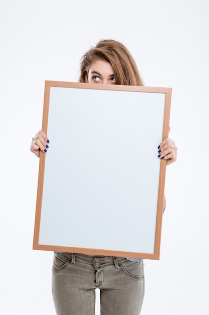 Portrait d'une jeune femme peeping over blank board isolé sur fond blanc