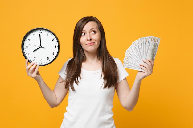 Portrait d'une jeune femme perplexe dans des vêtements décontractés blancs tenant une horloge ronde, fan d'argent liquide en billets en dollars isolés sur fond orange jaune. Concept de style de vie des gens. Maquette de l'espace de copie.