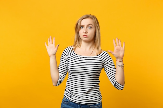 Portrait d'une jeune femme perplexe en vêtements rayés levant les mains, montrant des paumes isolées sur fond de mur orange jaune en studio. Concept de mode de vie des émotions sincères des gens. Maquette de l'espace de copie.