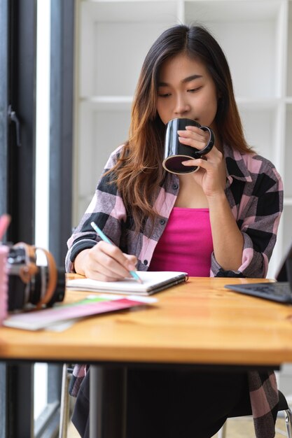 Portrait de jeune femme pigiste buvant une pause-café tout en écrivant sur un cahier vierge au café