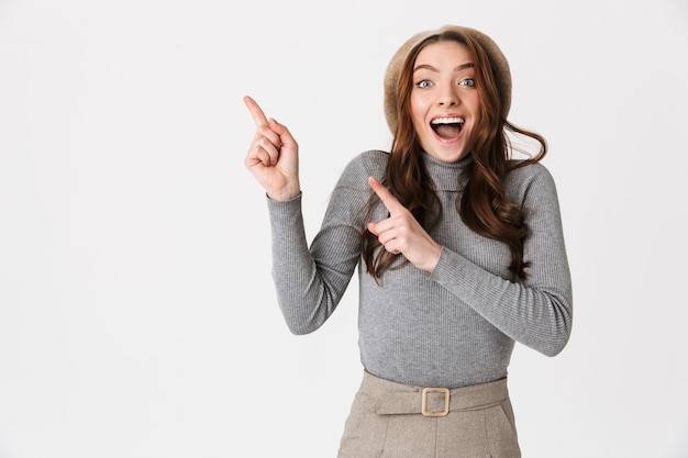 Portrait de jeune femme portant un chapeau souriant et pointant les doigts de côté au fond isolé sur mur blanc