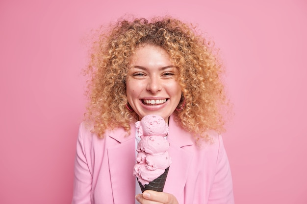 Portrait d'une jeune femme positive aux cheveux bouclés sourit joyeusement a la bonne humeur mange un dessert savoureux aime la crème glacée glacée froide habillée formellement isolée sur un mur rose. Délicieux goûter d'été
