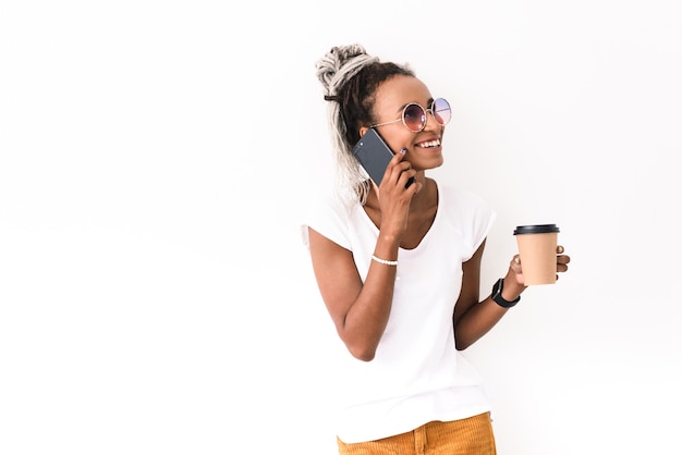 Portrait d'une jeune femme positive souriante heureuse avec des dreads posant isolé sur blanc parler par téléphone mobile, boire du café.