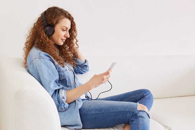 Portrait d'une jeune femme ravie écoute sa musique préférée via un téléphone intelligent moderne et des écouteurs, vêtue de vêtements en jean à la mode, connectée à Internet sans fil.