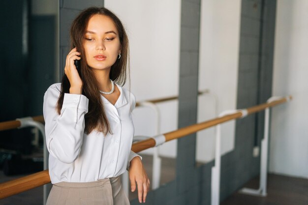 Portrait d'une jeune femme réfléchie dans des vêtements élégants parlant sur un téléphone portable debout dans la chambre avec un miroir regardant vers le bas