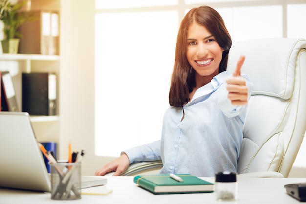 Portrait d'une jeune femme réussie assise au bureau avec le pouce levé et regardant la caméra avec le sourire sur son visage.