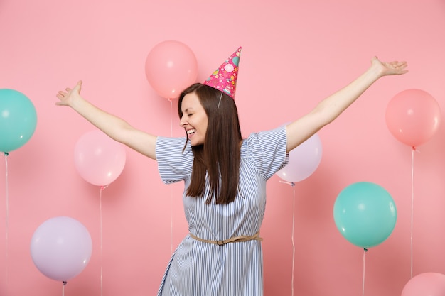 Portrait d'une jeune femme riante aux yeux fermés en chapeau d'anniversaire et robe bleue écartant les mains sur fond rose avec des ballons à air colorés. Fête d'anniversaire, concept d'émotions sincères.