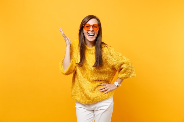 Portrait de jeune femme riante en pull de fourrure, pantalon blanc, lunettes orange coeur écartant les mains isolées sur fond jaune vif. Les gens émotions sincères, concept de style de vie. Espace publicitaire.