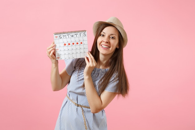 Portrait de jeune femme en robe bleue, chapeau tenant le calendrier des périodes pour vérifier les jours de menstruation isolés sur fond rose tendance brillant. Concept médical, sanitaire, gynécologique. Espace de copie.
