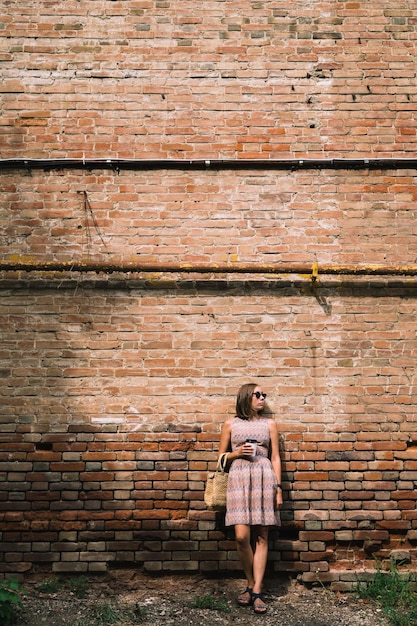 Portrait de jeune femme en robe et lunettes de soleil s'appuyant sur un mur de briques avec une tasse de café