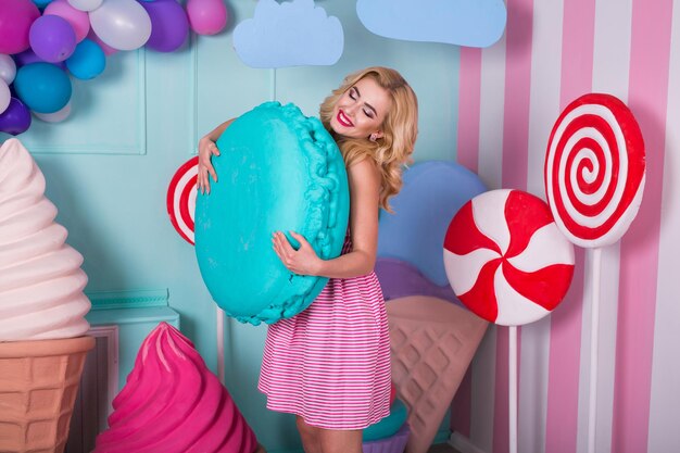 Portrait de jeune femme en robe rose tenant un gros macaron et posant sur fond décoré