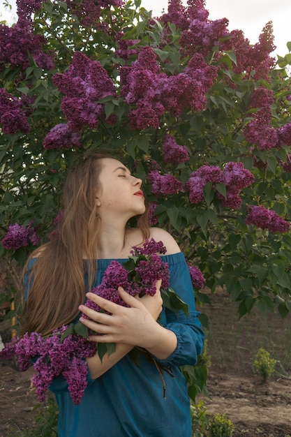 Portrait de jeune femme romantique en gros plan dans un jardin de lilas en fleurs Histoire de printemps La femme aux cheveux bruns sourit avec une fleur dans les cheveux en fermant les yeux et en profitant de l'ambiance printanière