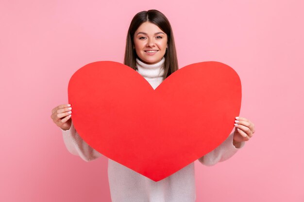 Portrait de jeune femme romantique tenant un grand cœur rouge, exprimant l'amour, célébrant la Saint-Valentin, portant un pull blanc de style décontracté. Tourné en studio intérieur isolé sur fond rose.