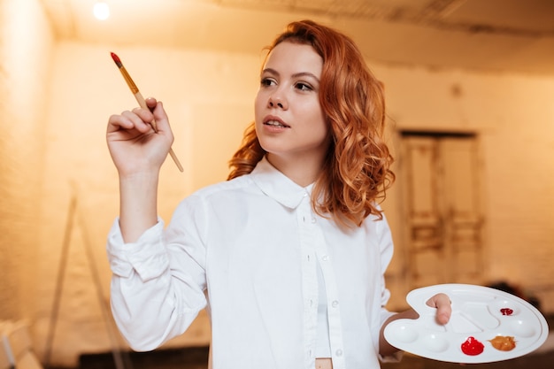 Portrait de jeune femme rousse concentrée peintre avec peintures à l'huile et peinture à la palette.