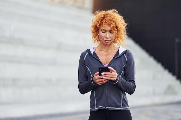 Portrait de jeune femme rousse européenne qui se tient debout avec un téléphone dans les mains à l'extérieur