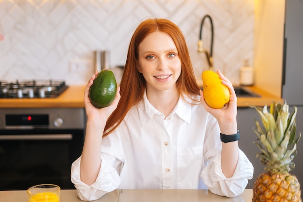 Portrait de jeune femme rousse gaie attrayante tenant dans les mains la mangue et l'avocat assis à