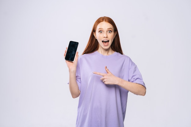 Portrait d'une jeune femme rousse heureuse pointant du doigt un téléphone portable à écran blanc