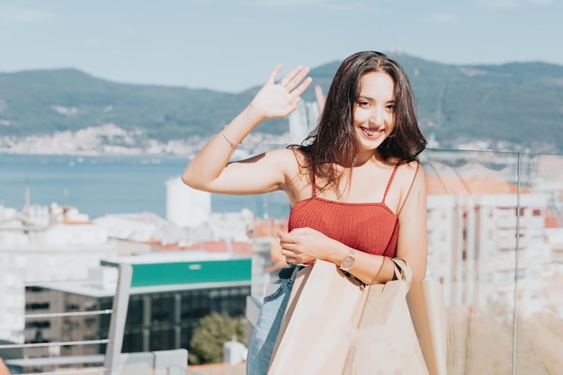 Portrait d'une jeune femme saluant l'acheteur à l'appareil photo heureux lors de l'achat et du transport de sacs à provisions. Espace de copie, concept de magasinage actif. Le consumérisme et le réseau social des jeunes.