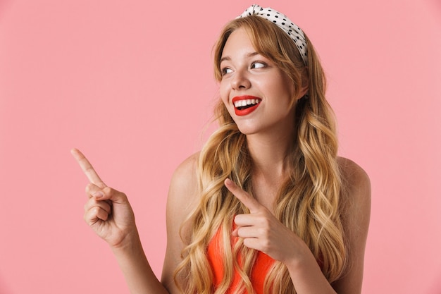 Portrait d'une jeune femme séduisante aux longs cheveux bouclés souriante et pointant du doigt le fond isolé sur un mur rose