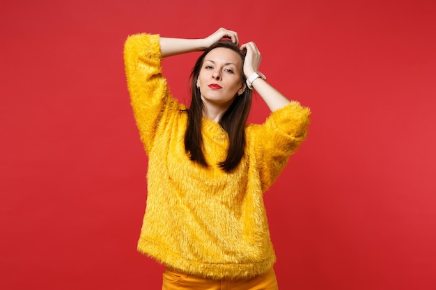 Portrait d'une jeune femme séduisante en pull de fourrure jaune debout avec les mains près de la tête isolée sur fond de mur rouge vif en studio. Les gens émotions sincères, concept de style de vie. Maquette de l'espace de copie.