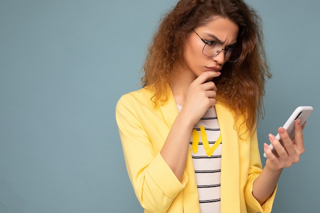 Portrait de jeune femme séduisante sérieuse réfléchie avec des cheveux blonds foncés bouclés portant une veste jaune et des lunettes optiques isolées sur le mur tenant et à l'aide de téléphone en regardant l'écran mobile.