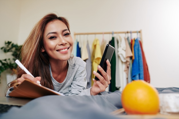 Portrait de jeune femme séduisante souriante allongée sur le lit, vérifiant son téléphone et prenant des notes dans le planificateur