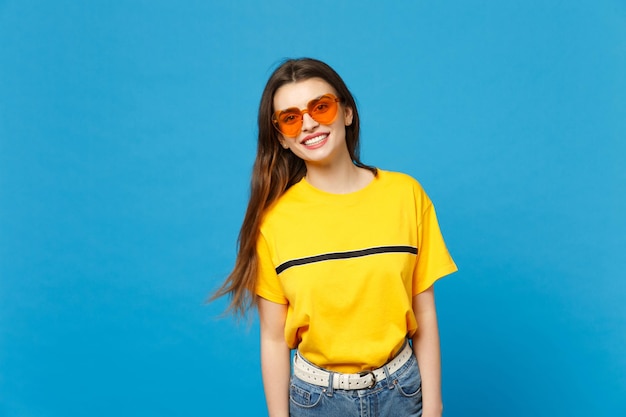 Portrait de jeune femme séduisante souriante dans des vêtements décontractés vifs, lunettes de coeur debout, regardant la caméra isolée sur fond de mur bleu en studio. Concept de style de vie des gens. Maquette de l'espace de copie.