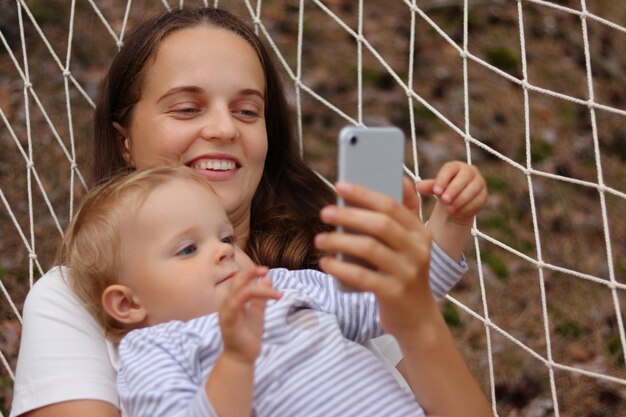 Portrait d'une jeune femme séduisante souriante avec sa fille mère et enfant se relaxant dans un hamac dans la forêt et utilisant un téléphone intelligent regardant l'écran de l'appareil en regardant des dessins animés en ligne ensemble
