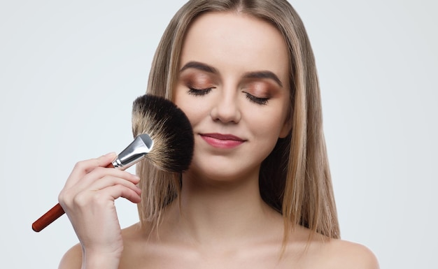 Portrait d'une jeune femme séduisante tenant un pinceau à la main tout en appliquant du maquillage sur fond gris