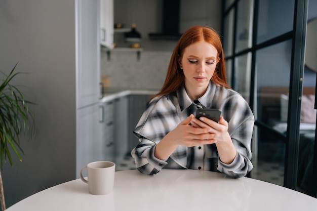 Portrait d'une jeune femme sérieuse et concentrée assise à une table partageant des nouvelles sur les réseaux sociaux via un smartphone regardant l'écran du téléphone Jolie femme rousse lisant du texte