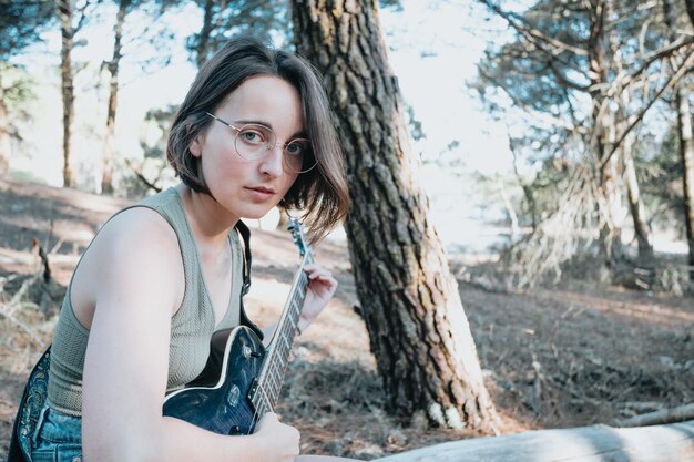 Portrait d'une jeune femme sérieuse hipster tenant la guitare à l'extérieur du parc forestier de la ville. En s'amusant à apprendre une nouvelle compétence, la musique joue un style saisonnier. Jeune fille aux cheveux courts. Espace de copie