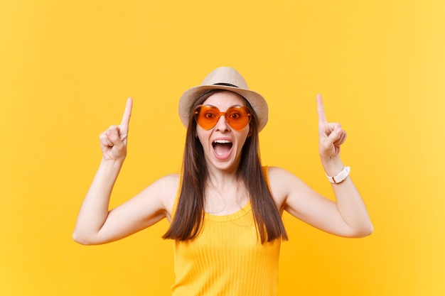 Portrait de jeune femme souriante au chapeau d'été de paille, lunettes orange pointant l'index vers le haut sur l'espace de copie isolé sur fond jaune. Les gens émotions sincères, concept de style de vie. Espace publicitaire.